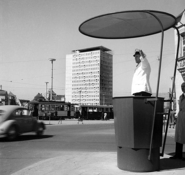 Plärrer mit Hochhaus 1953: Der Autoverkehr wurde lange Zeit an den wichtigen Verkehrsknotenpunkten von einem Polizisten geregelt. Im Hintergrund der Szene ist das Plärrer-Hochhaus zu sehen, das als städtebauliche Sensation galt. Das 1951-53 von Wilhelm Schlegtendal (1906-1994) für die Städtischen Werke errichtete Gebäude war Bayerns erstes Hochhaus. Seine elegante Wirkung erzielt das 55 Meter hohe Bürohaus durch die sich ab dem 5. Stock jeweils um einen Zentimeter verjüngenden Geschosse. Foto: © Hochbauamt