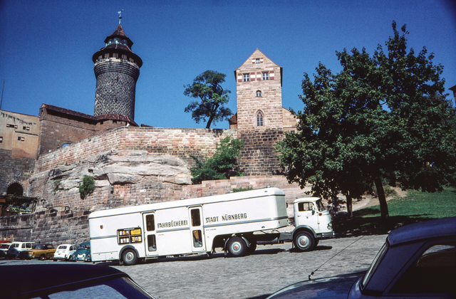 Der Bücherbus der Stadtbibliothek überquert den Ölberg. Seit 1959 werden zusätzlich zu den stationären Außenstellen der Stadtbib-liothek mobile Standorte durch die Fahrzeuge der Fahrbibliothek versorgt. Im Hintergrund, auf dem Gipfel des Burgbergs aus rötli-chem Sandstein, links der Sinwellturm und rechts die Giebelfassade der Walpurgiskapelle. | Foto: Hochbauamt, 1961. (StadtAN A 55 Nr. I-10-5-2)