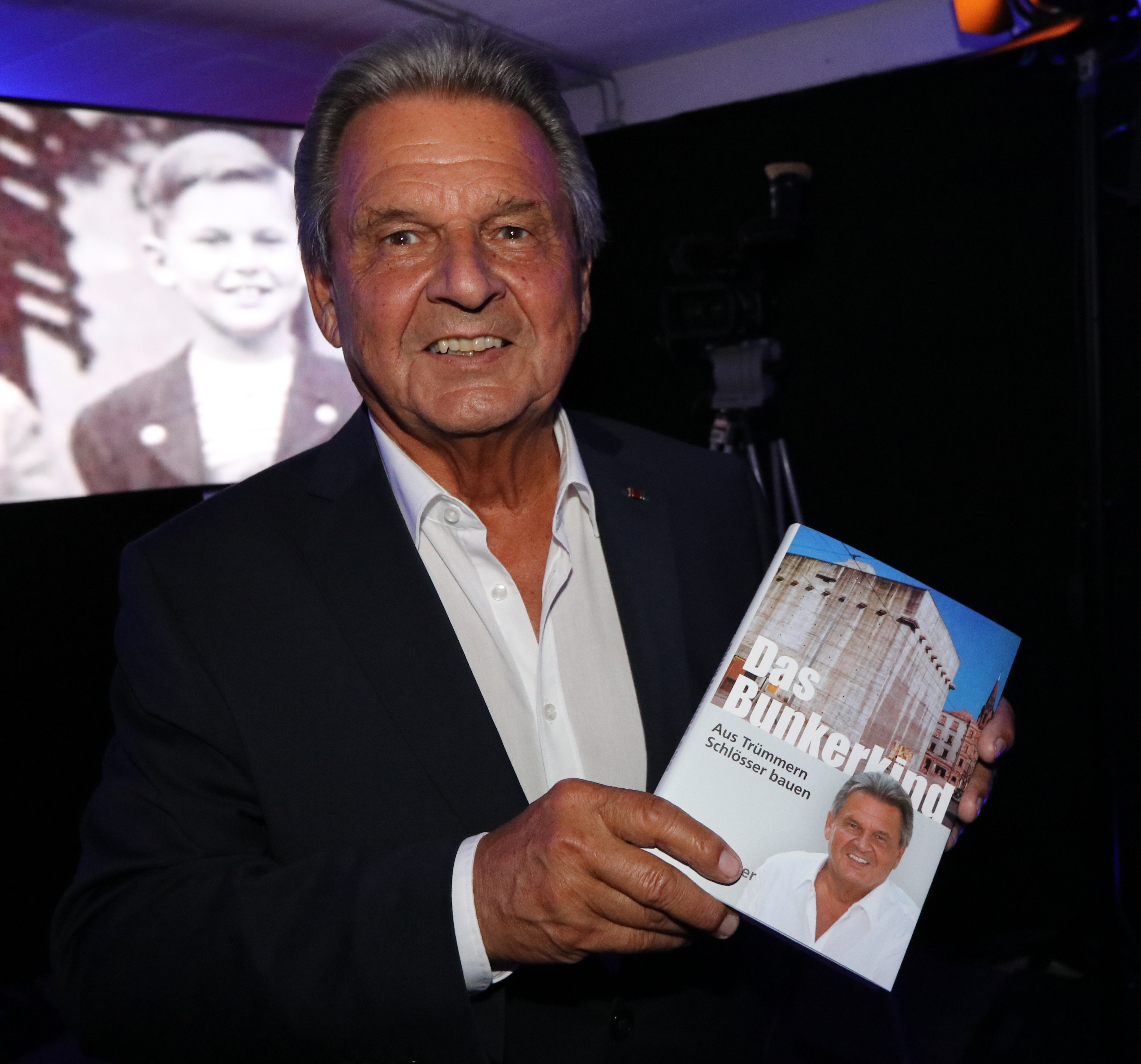 DEFACTO X-Gründer Erich Schuster präsentiert sein Buch ,,Das Bunkerkind“. Im Hintergrund Erich Schuster als Junge.