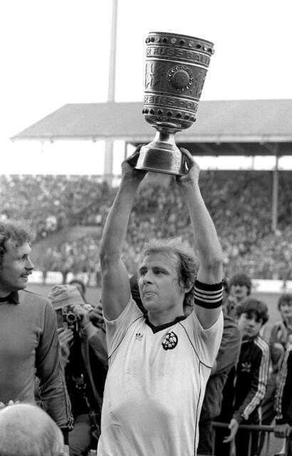 Stuttgart: Der Frankfurter Eintracht-Stürmer und Kapitän Bernd Hölzenbein (M) reckt an der Seite von Torhüter Jürgen Pahl (l) den eroberten DFB-Pokal triumphierend hoch. Eintracht Frankfurt gewann vor 71.000 Zuschauern im Stuttgarter Neckarstadion das DFB-Pokalendspiel gegen den 1. FC Kaiserslautern mit 3:1 und sicherte sich den Cup damit zum drittenmal. Hölzenbein ist mit 78 Jahren gestorben.  | Foto: picture alliance / dpa 
